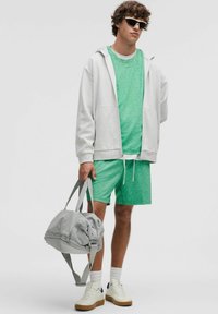 Graue Zip-Up-Hoodie über einem grünen T-Shirt mit kurzen Ärmeln und passenden grünen Shorts. Hält eine graue Sporttasche mit grauen Trägern. Weiße Turnschuhe.