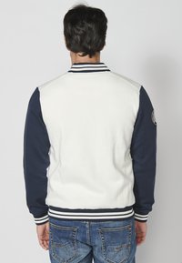 Veste de style universitaire en mélange de coton blanc et marine. Dotée d'un col côtelé, de poignets rayés, et d'un patch logo sur la manche gauche.
