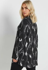 Long black and white patterned jacket with a relaxed fit, ribbed texture, and side slits, worn over dark pants.