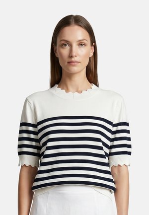 Vrouw met steil bruin haar die een wit shirt met korte mouwen draagt met zwarte horizontale strepen en gegolfde randen aan mouwen en hals.