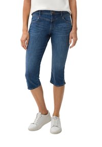 Dunkelblaue Denim-Caprihosen mit tailliertem Schnitt, umgeschlagenem Saum, fünf Taschen und bronzenen Beschlägen. Kombiniert mit weißen Sneakers.