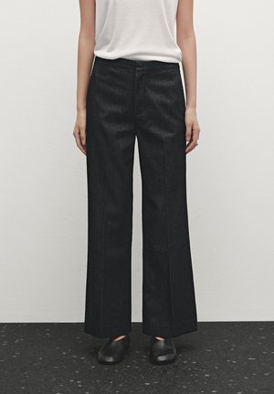 Kobieta ubrana w czarne spodnie o szerokich nogawkach, biały bezrękawnik i czarne buty, stoi na tle jasnej, jednolitej ściany.