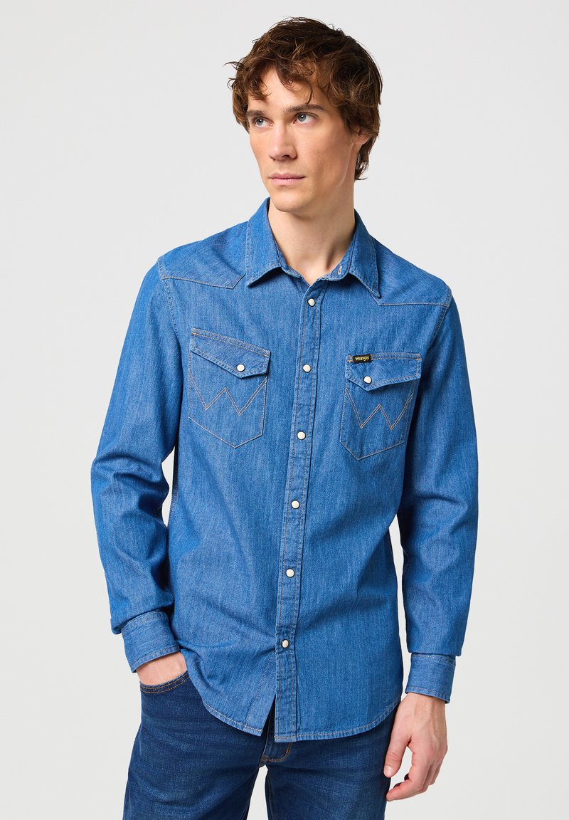 Camisa de mezclilla de manga larga en azul claro. Cuenta con dos bolsillos en el pecho con solapas puntiagudas, cierres de botón y costuras en contraste. Textura suave.