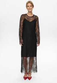 Černé krajkové šaty s dlouhými rukávy, průhlednou texturou a výstřihem do V. Má volný siluetu a vrstvenou sukni, doplněné červenými podpatky.