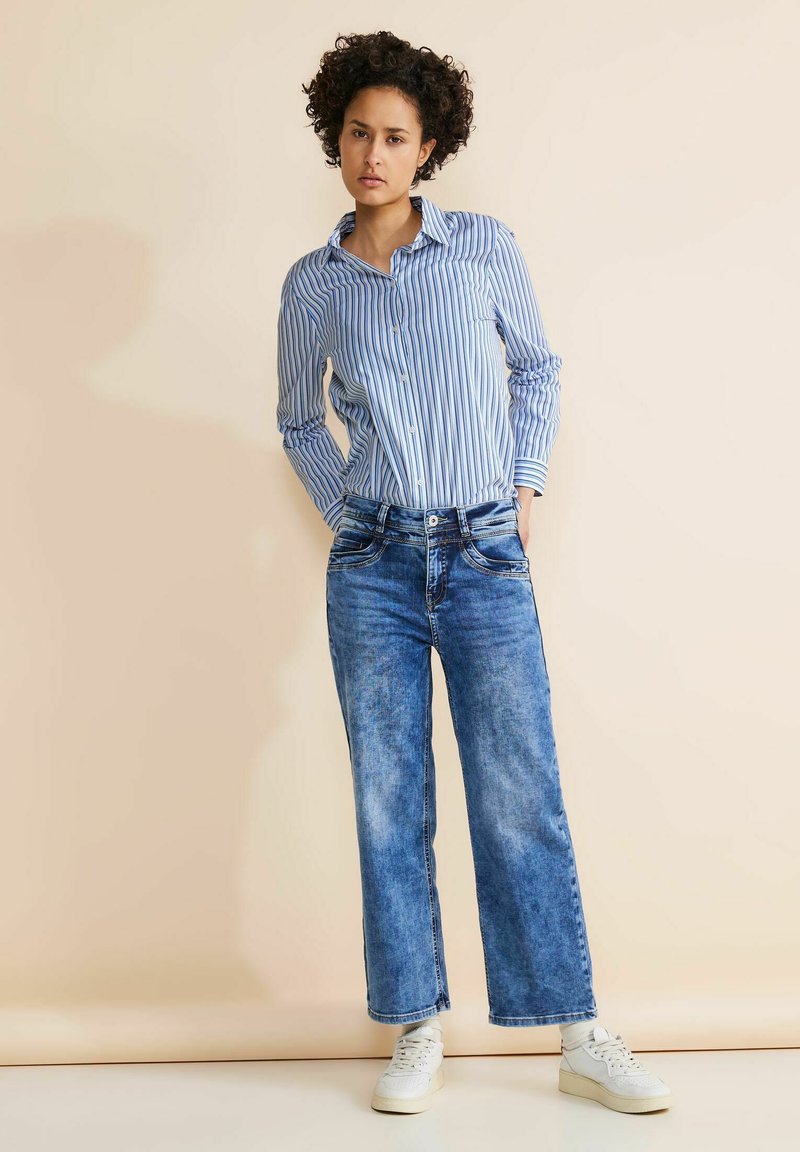 Randig blå och vit skjorta med knappar, avslappnat passande blekta blå jeans och vita sneakers. Modellen står med händerna i fickorna mot en ljus bakgrund.
