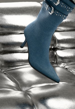Stivaletto in denim blu con punta affusolata, tacco piccolo e dettagli in fibbia argentata, poggiato su una superficie metallica lucida.