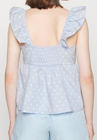 Woman wearing a light blue sleeveless top with white polka dots and ruffled straps, paired with light blue shorts, facing away.