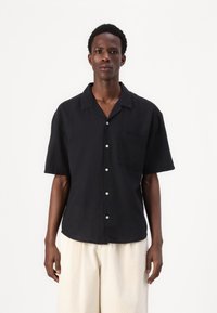 Camisa negra de manga corta hecha de lino, con un cierre frontal de botones, un bolsillo en el pecho y un corte relajado. Textura lisa, sin patrones.