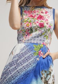 Kukkakuosinen mekko sinisävyisellä liukuvärillä. Mekossa on erilaisia printtejä, hihaton malli ja koristeelliset yksityiskohdat pääntiessä.