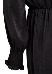 Camisa negra de manga larga con puños fruncidos elásticos y borde con volantes, cintura parcialmente visible con detalle elástico en tela ligera.