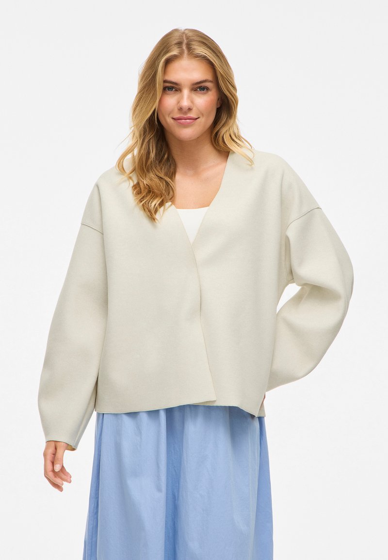 Šviesiai bežinė apvyniojama svetrė su plačiomis rankovėmis ir gilia V formos iškirpte. Minkšta medžiaga su minimalizuotu dizainu. Derinama su šviesiai mėlyna plazdančia sijonu.