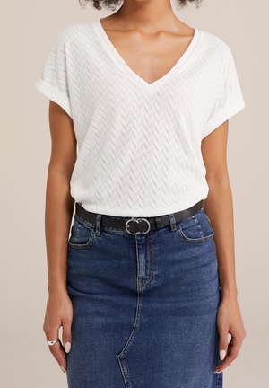 Vrouw draagt een wit V-hals kort mouw textuur top, ingestopt in een hooggetailleerde blauwe denim rok met een zwarte riem en zilveren gesp.