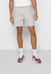 Light gray checkered swim shorts with an elastic waist and drawstring, featuring an embroidered orange logo. Paired with red and blue sneakers.
