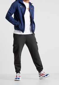 Navy windbreaker with a hood, white t-shirt, dark gray cargo pants, and multicolored athletic shoes. Features prominent logo accents.