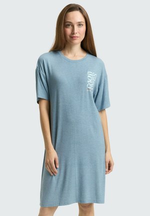 Vrouw met steil bruin haar, gekleed in een loszittende lichtblauwe T-shirtjurk met witte verticale tekst op de borst.