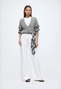 Mujer que lleva una chaqueta gris de lana, camisa blanca, pantalones anchos blancos con una bufanda en blanco y negro atada al cinturón y sandalias negras.