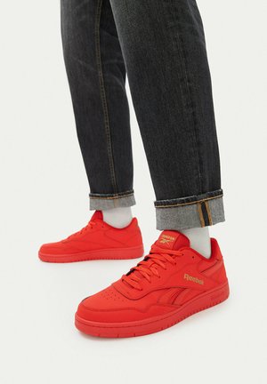 Person trägt leuchtend rote Reebok-Sneakers mit weißen Socken und schwarzen umgeschlagenen Jeans und steht vor einem schlichten weißen Hintergrund.