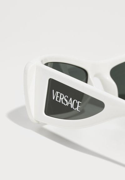 Lunettes de soleil blanches avec des verres foncés, arborant un design contemporain. Le nom de la marque "Versace" est inscrit sur le côté en lettres majuscules.