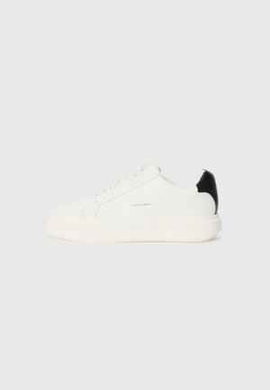 White leather sneaker with a black accent on the heel, featuring round toe, flat laces, and a thick white sole. Calvin Klein logo displayed.
