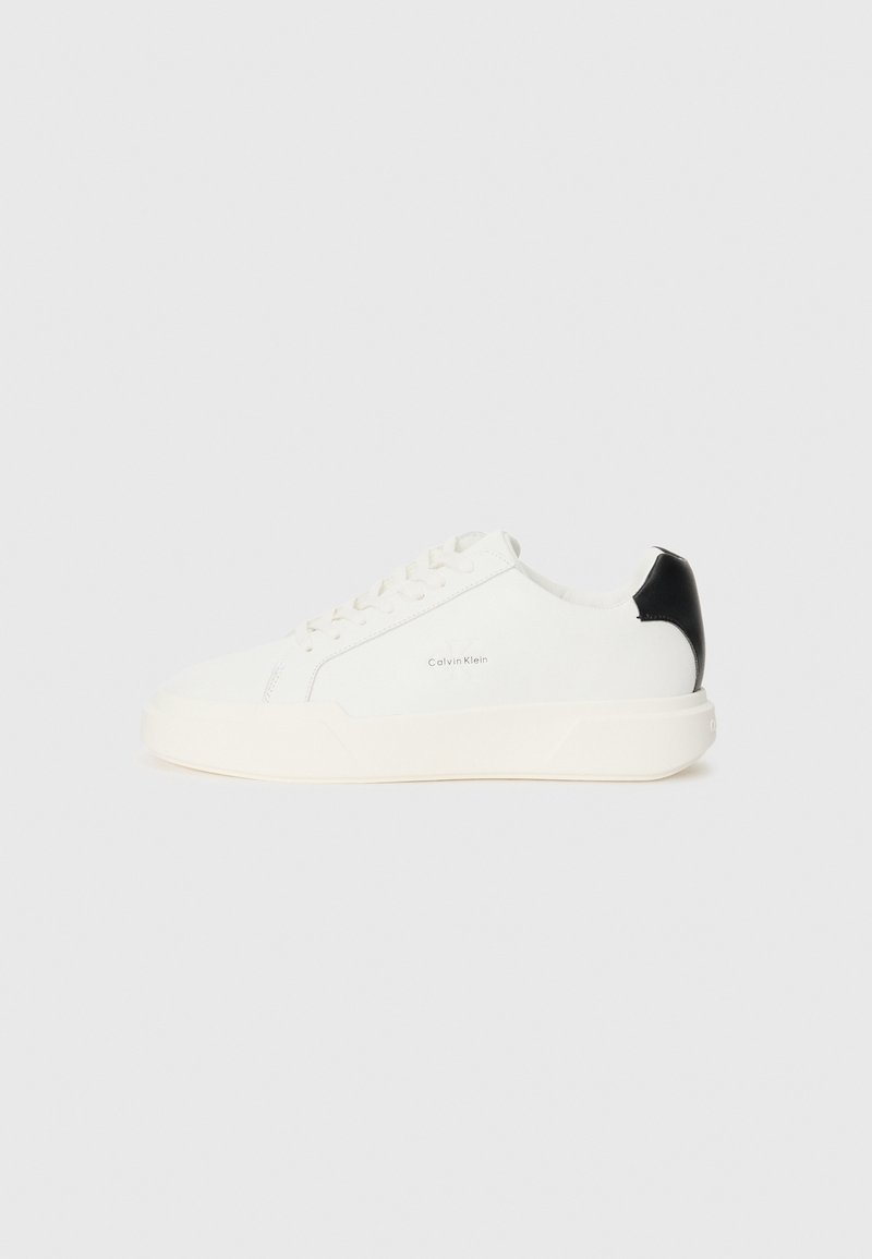 White leather sneaker with a black accent on the heel, featuring round toe, flat laces, and a thick white sole. Calvin Klein logo displayed.