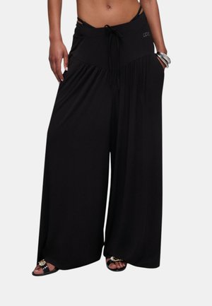 Donna che indossa pantaloni neri a gamba larga con coulisse in vita e sandali neri, in piedi con le mani nelle tasche.