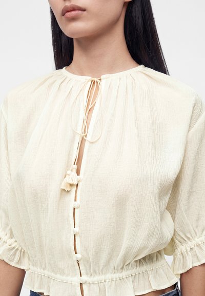 Femme portant un chemisier texturé de couleur crème avec des boutons sur le devant et un lien avec des pompons au niveau de l'encolure.