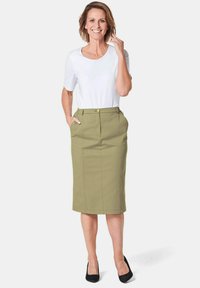 Olivgrüner Bleistiftrock mit vorderen Falten und Taschen, kombiniert mit einem schmal geschnittenen weißen T-Shirt. Schwarze High Heels runden das Outfit ab.
