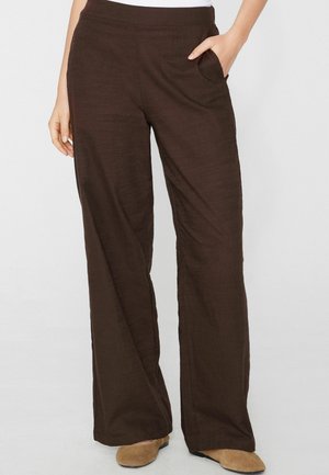 Kobieta nosząca luźne, ciemnobrązowe spodnie z bocznymi kieszeniami oraz jasnobrązowe, płaskie buty na jasnym tle.