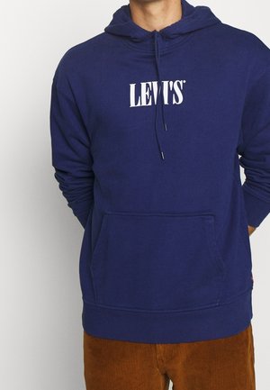 Homme portant un sweat à capuche Levi's bleu avec poche avant et cordons, associé à un pantalon en velours côtelé marron, debout devant un fond blanc.