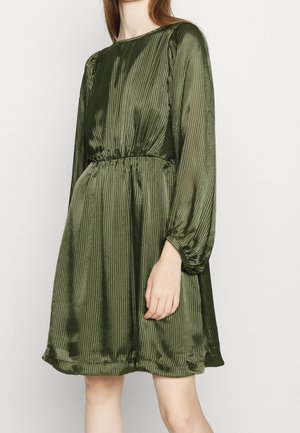 Kobieta w oliwkowej plisowanej sukience o długości do kolan, z długim rękawem i wcięciem w talii, na jasnym tle.