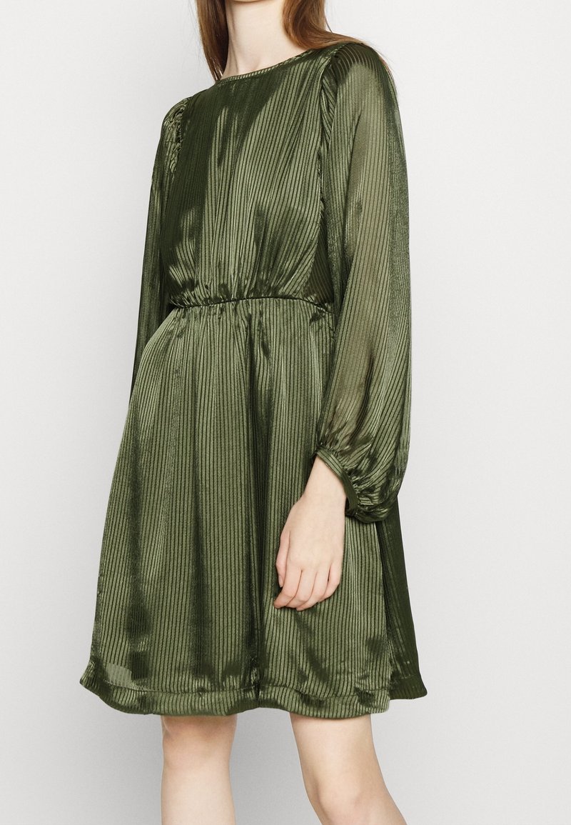 Femme portant une robe plissée vert olive mi-longue à manches longues avec une taille cintrée sur un fond clair.
