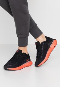 Chaussures de sport noires avec une tige synthétique texturée, présentant des accents orange sur la semelle et un design flexible et rembourré.