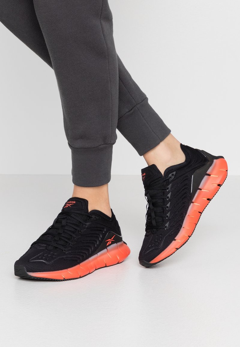 Chaussures de sport noires avec une tige synthétique texturée, présentant des accents orange sur la semelle et un design flexible et rembourré.