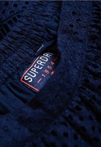 Tkanina w kolorze granatowym z teksturowym wzorem i marszczeniami. Posiada widoczną metkę z napisem "SUPERDRY" oraz kontrastowe pomarańczowe akcenty.