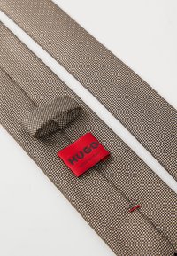 Barna textil nyakkendő apró szőtt mintázattal, piros "HUGO" címkével. Sima textúrájú, hegyes végű, és rögzítő hurkával rendelkezik.