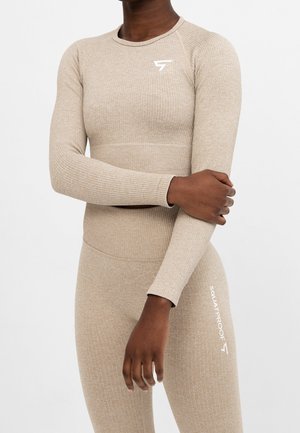 Persoon draagt een beige, geribbelde, lange mouwen sporttop en bijpassende legging met het logo "SQUATPROOF" op de dij, armen gekruist.