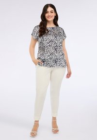 Tummansininen ja valkoinen lehvämönkijä, löysällä istuvuudella, yhdistettynä kermaisiin tapered-housuihin. Malli seisoo kädet taskuissa, jalassaan nauhalla varustetut korot.