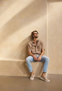 Hnědá košile s krátkým rukávem, světle modré dobře padnoucí džíny a béžové slip-on boty; sedící opřený o texturovanou béžovou zeď se měkkým osvětlením.