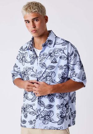 Jeune homme aux cheveux blonds courts portant une chemise à boutons bleu clair à motifs floraux, manches retroussées, debout devant un fond uni.