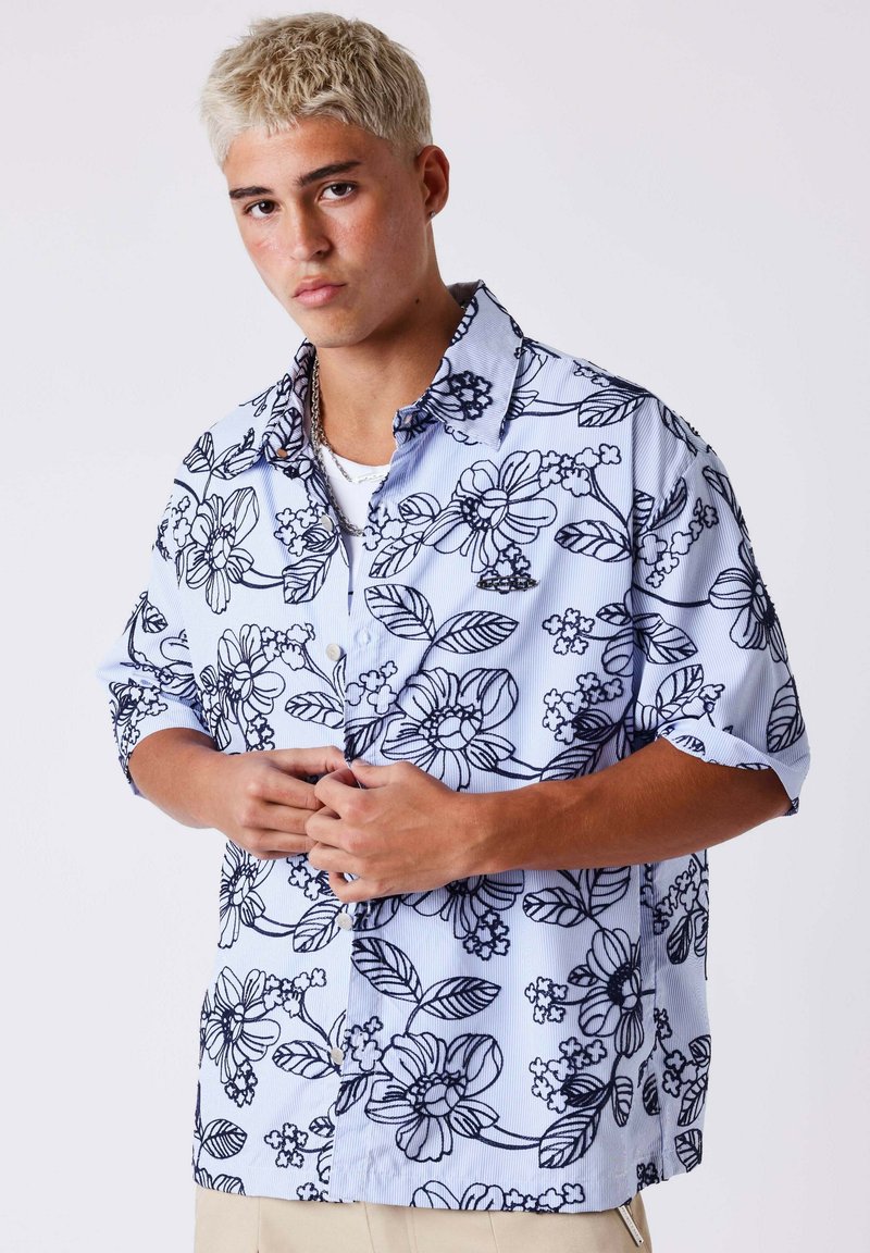 Jeune homme aux cheveux blonds courts portant une chemise à boutons bleu clair à motifs floraux, manches retroussées, debout devant un fond uni.
