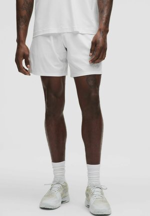 Mężczyzna ubrany w białe spodenki sportowe, białe skarpetki i białe buty do biegania, stojący na jednolitym jasnym tle.