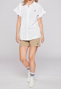 Vit skjorta med volangärmar och liten broderad logotyp, tillsammans med beige shorts med en matchande logotyp. Vita sneakers och randiga strumpor.