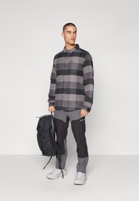 Svart og grå rutete flanellskjorte med knapper, brukt over grå og svarte turbukser. Personen holder en svart ryggsekk med stropper.