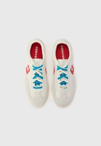 Baskets blanches avec une pointe en daim texturé et un dessus lisse. Présentent des lacets bleus, un intérieur rouge et un logo en forme d'étoile rose sur le côté.