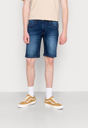 Mand iført falmede blå denimshorts, beige t-shirt, hvide crew-sokker og sennepsgule sneakers stående foran en ensfarvet baggrund.