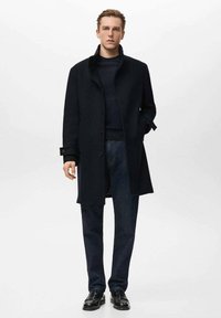 Ciemnogranatowy wełniany płaszcz z wysokim kołnierzem, jednorzędowy, z bocznymi kieszeniami. W zestawie z ciemnymi dżinsami i skórzanymi butami.