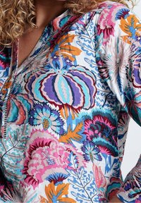 Blouse florale colorée présentant un mélange de motifs violets, bleus, orange et roses, avec un col à boutons et un tissu léger.