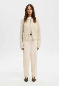 Femme debout, vue de face, portant un cardigan en maille beige, un pantalon ample taille haute assorti, des chaussures pointues marron et une ceinture.