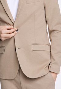 Beżowa marynarka z teksturowanej tkaniny, z pojedynczym czarnym guzikiem, dwiema przednimi kieszeniami i dopasowanym krojem z wyłogami.