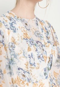 Blommig blus i lätt, genomskinlig tyg med blå och beige blommor. Inkluderar delikat spetskant och veckad design.